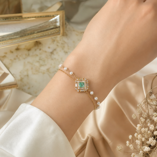 A delicate gold bracelet with a central green emerald-cut stone and small white pearls, shown worn on a person's wrist against a soft beige satin and marble background.