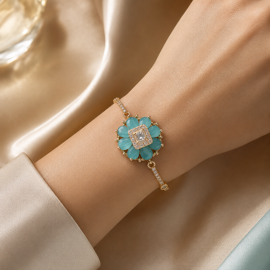 A gold-tone flower-shaped bracelet with turquoise blue stones and a central clear crystal, shown worn on a person's wrist against a soft beige satin background.