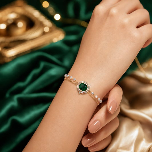 A close-up of a woman's wrist wearing a delicate gold bracelet with a central green oval stone and small white pearls, set against a background of dark green satin fabric and gold drapery.
