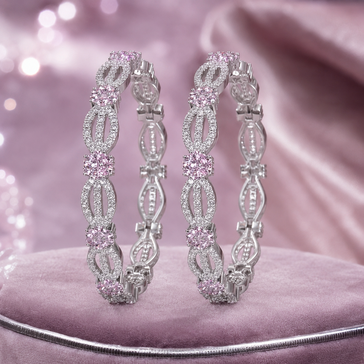 A pair of silver-toned huggie bangles decorated with pink gemstones and clear crystals, displayed on a pink velvet surface with a matching pink satin background.