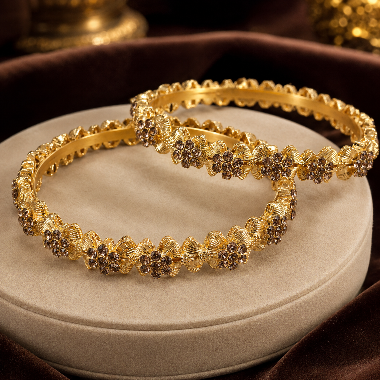 Two ornate gold bangles with floral rhinestone embellishments, displayed on a round beige cushion with dark brown velvet fabric in the background.