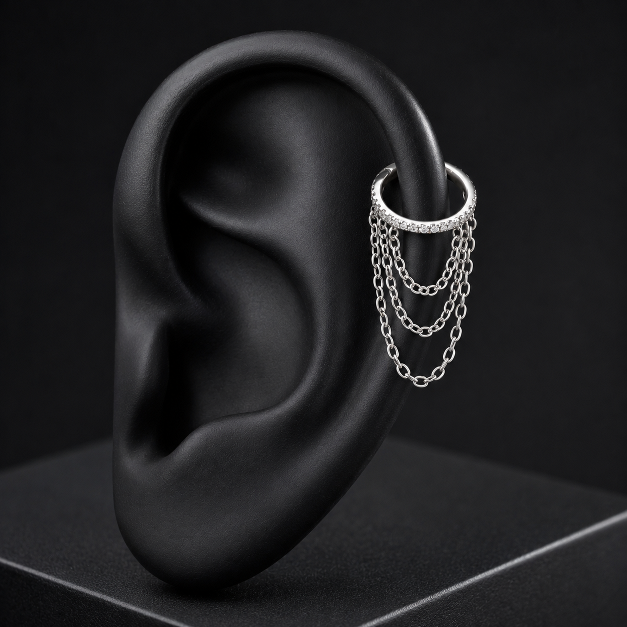 A silver ear cuff with three draped chain layers, displayed on a matte black ear stand against a dark background.