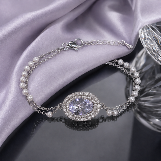 A silver chain bracelet with white pearls and a central oval crystal pendant surrounded by a halo of small clear stones, displayed on draped lavender satin fabric over a dark reflective surface.