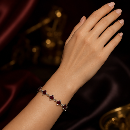 A delicate silver-toned bracelet with alternating square-cut deep red gemstones and small clear stones, shown worn on a woman's wrist against a dark, elegant background with draped burgundy fabric and soft gold bokeh.