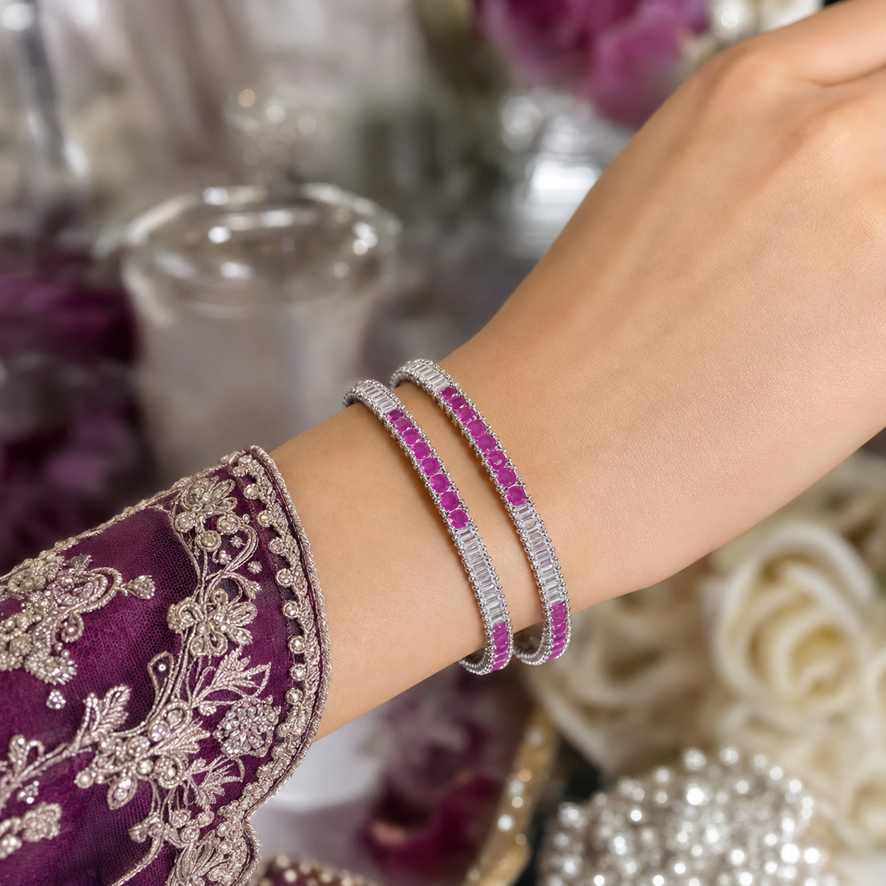 A close-up of a person's wrist wearing two silver-toned bangles adorned with bright pink gemstones and clear stones, with a purple embroidered sleeve visible at the side.