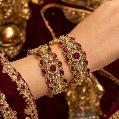 A close-up of a person's wrist wearing ornate gold bangles decorated with red stones and white faux pearls, set against a rich maroon and gold traditional outfit background.