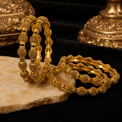 A set of gold bangles with textured, beaded spherical details, displayed on a light stone slab against a black velvet background with ornate gold-toned decorative objects.