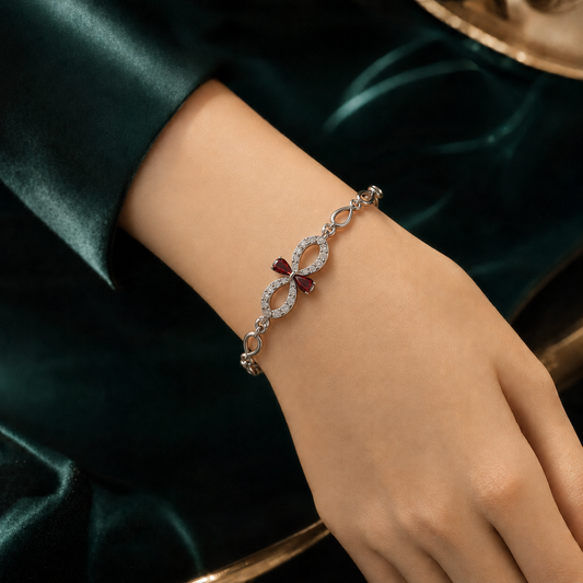 A close-up of a person's wrist wearing a delicate silver-toned infinity bracelet adorned with small clear stones and a central bow motif featuring deep red gemstones, set against a dark green satin fabric background.