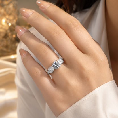 A close-up of a woman's hand wearing a silver-toned diamond ring with a central round-cut stone and smaller side stones, set against a soft white blouse and warm blurred background.