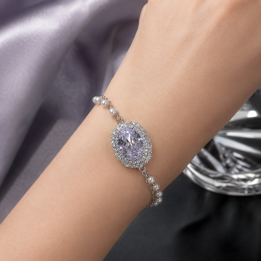 A close-up of a silver-toned bracelet with a large oval lavender gemstone surrounded by a halo of small clear stones, worn on a person's wrist. The bracelet also features delicate pearl-like links.