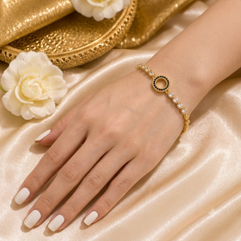 A gold bracelet with a circular charm featuring black and white stones, worn on a woman's wrist. The hand and wrist are resting on a beige satin fabric, with white flowers and a gold textured plate in the background.