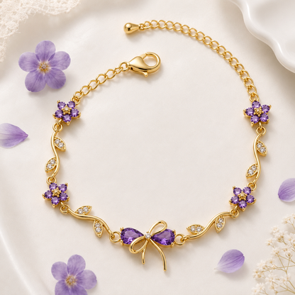 Gold chain bracelet with purple floral and bow charms, accented with small clear stones, displayed on a white surface with scattered purple flower petals.