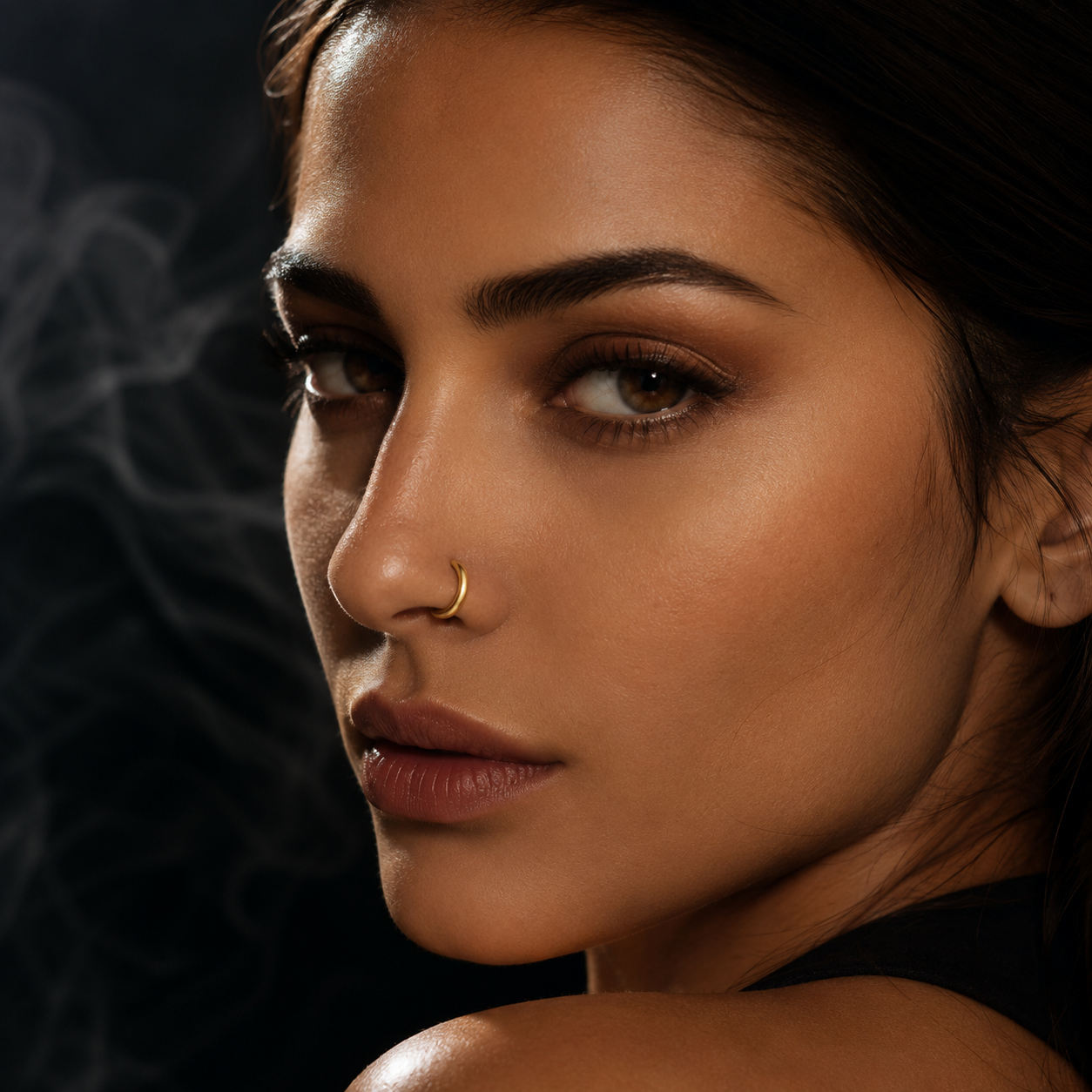 A close-up portrait of a woman wearing a small gold crescent moon nose ring against a dark smoky background.