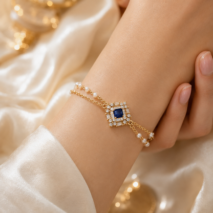 A gold double-strand bracelet with a central square blue gemstone surrounded by clear crystals, accented with small white pearls, shown worn on a person's wrist against a soft cream satin background.