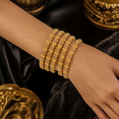 A close-up of a person's wrist wearing five stacked gold bangles adorned with small clear stones and bead details, set against a dark draped fabric background with ornate gold decorative elements.