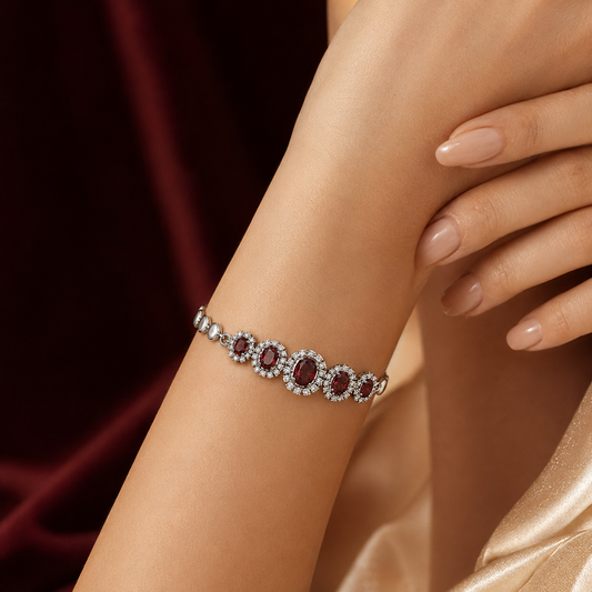 A close-up of a woman's wrist wearing a silver-toned bracelet with oval red gemstones and clear stones in a halo setting, against a dark red and gold satin background.