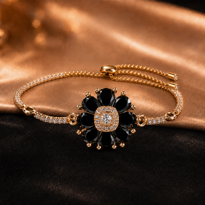 A gold-plated bracelet featuring a central flower motif with black gemstone petals, a clear central stone, and small clear accent stones along the band, displayed on a dark and gold satin background.