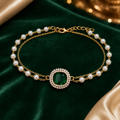 A gold chain necklace with white pearls and a central emerald-cut green gemstone surrounded by small clear stones, displayed on a dark green velvet fabric background.