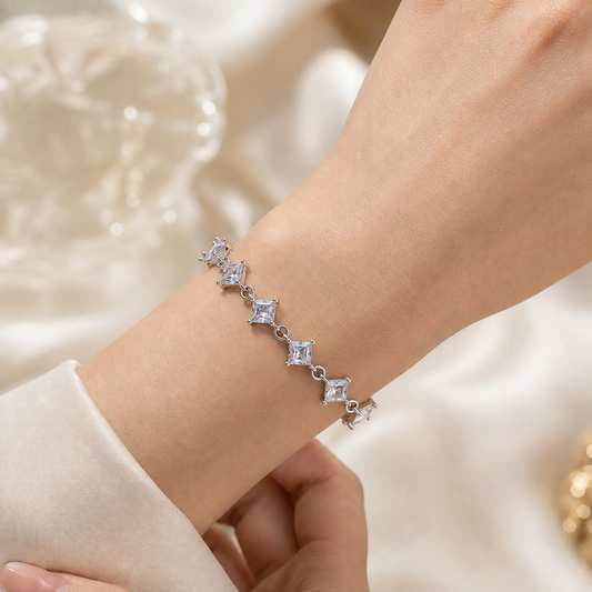 A close-up of a delicate silver bracelet with four star-shaped clear gemstones worn on a person's wrist, set against a soft cream-colored fabric background.
