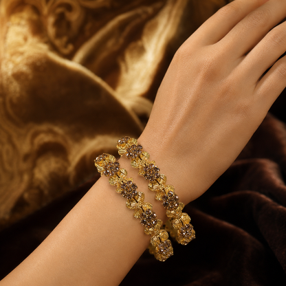 A close-up of a person's wrist wearing two matching gold-toned bangle bracelets adorned with floral clusters of clear stones and small brown accents, set against a luxurious brown velvet background.