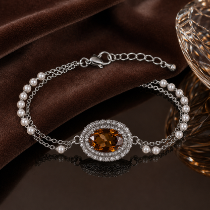 A silver chain bracelet with a central oval halo of clear stones and a warm amber-brown gemstone, accented by small white pearls, displayed on a dark brown velvet surface.
