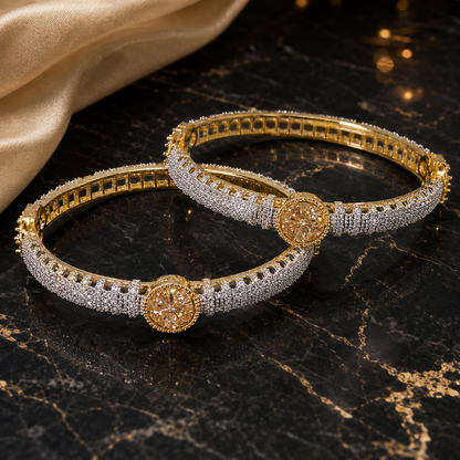 Two gold-plated bangle bracelets heavily embellished with clear pavé stones, featuring round gold-toned medallion centers, displayed on a black marble surface with a beige satin cloth in the background.