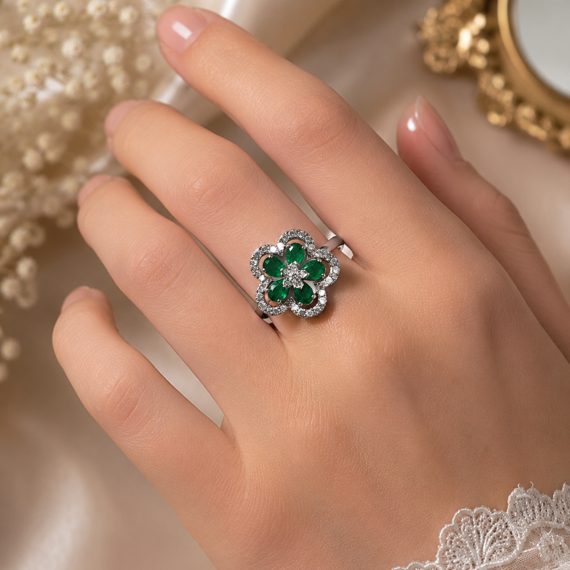 A silver-toned ring with a flower-shaped cluster of green emerald-cut stones and small clear accent stones, shown worn on a hand against a soft cream satin background with decorative pearls and lace.