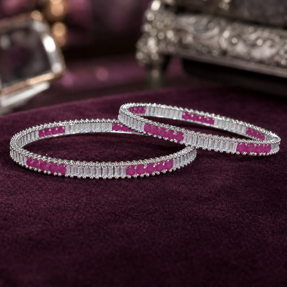 Two silver-toned bangles decorated with rectangular pink gemstones and clear stones, displayed on a dark purple velvet surface.