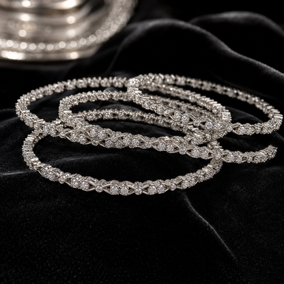 A stack of silver-toned tennis bracelets adorned with sparkling clear stones, arranged on a black velvet surface with a blurred silver tray in the background.