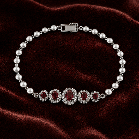 A pearl and silver bracelet featuring a central row of five oval red gemstones surrounded by clear crystals, displayed on a dark red velvet background.