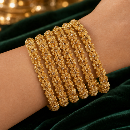 Five stacked gold bangles with intricate floral detailing and small clear stones, worn on a person's wrist against a dark green velvet background.