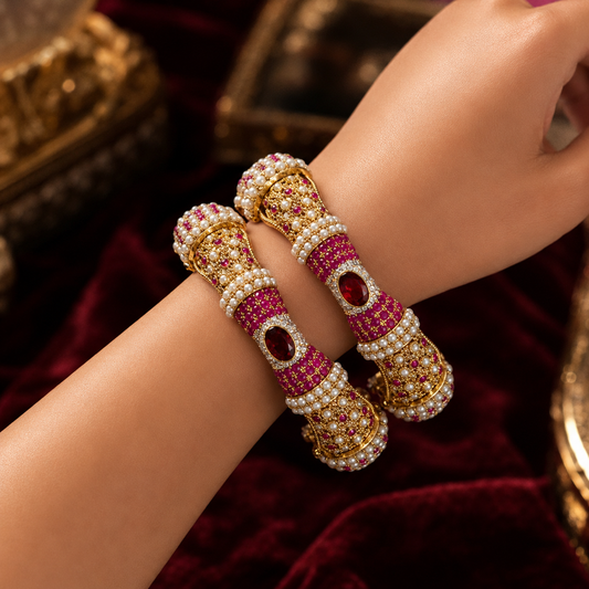A pair of ornate gold-plated bangles decorated with rows of white pearls and vibrant pink stones, featuring central red gemstone accents, shown worn on a person's wrist against a dark red velvet background.