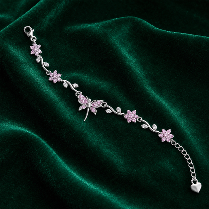 A silver-toned bracelet featuring pink flower and dragonfly motifs with small clear stones, displayed on a dark green velvet fabric background.