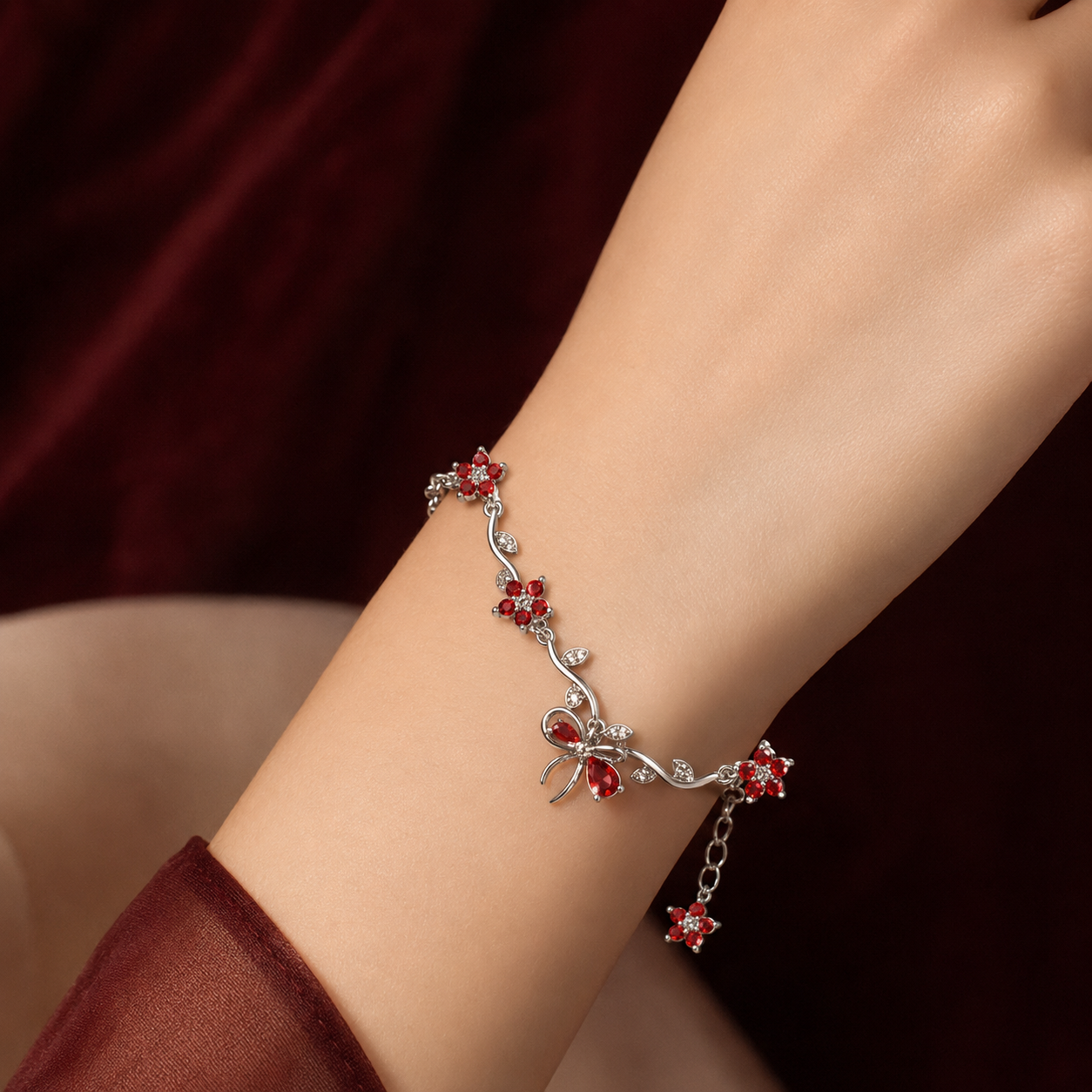 A delicate silver-toned bracelet with red crystal floral and bow motifs, shown worn on a person's wrist against a dark burgundy fabric background.