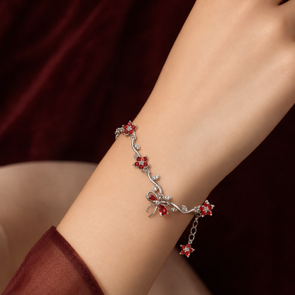 A delicate silver-toned bracelet with red crystal floral and bow motifs, shown worn on a person's wrist against a dark burgundy fabric background.