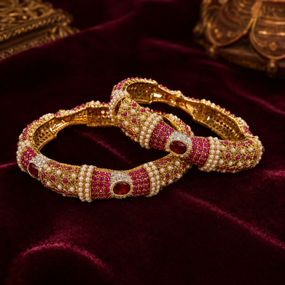 Two ornate gold-plated bangles decorated with rows of white pearls and red gemstones, displayed on a deep burgundy velvet surface.
