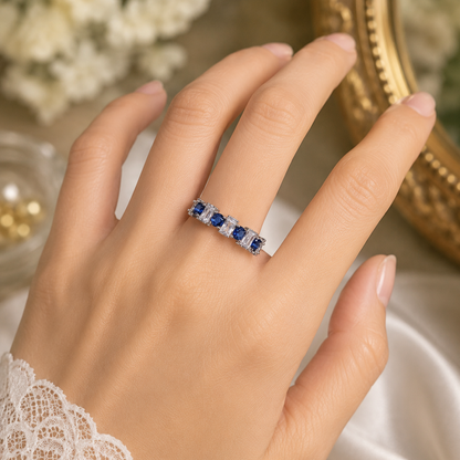 A close-up of a hand wearing a silver band ring set with five round-cut blue sapphires, surrounded by small clear stones, with a white lace cuff visible on the wrist.