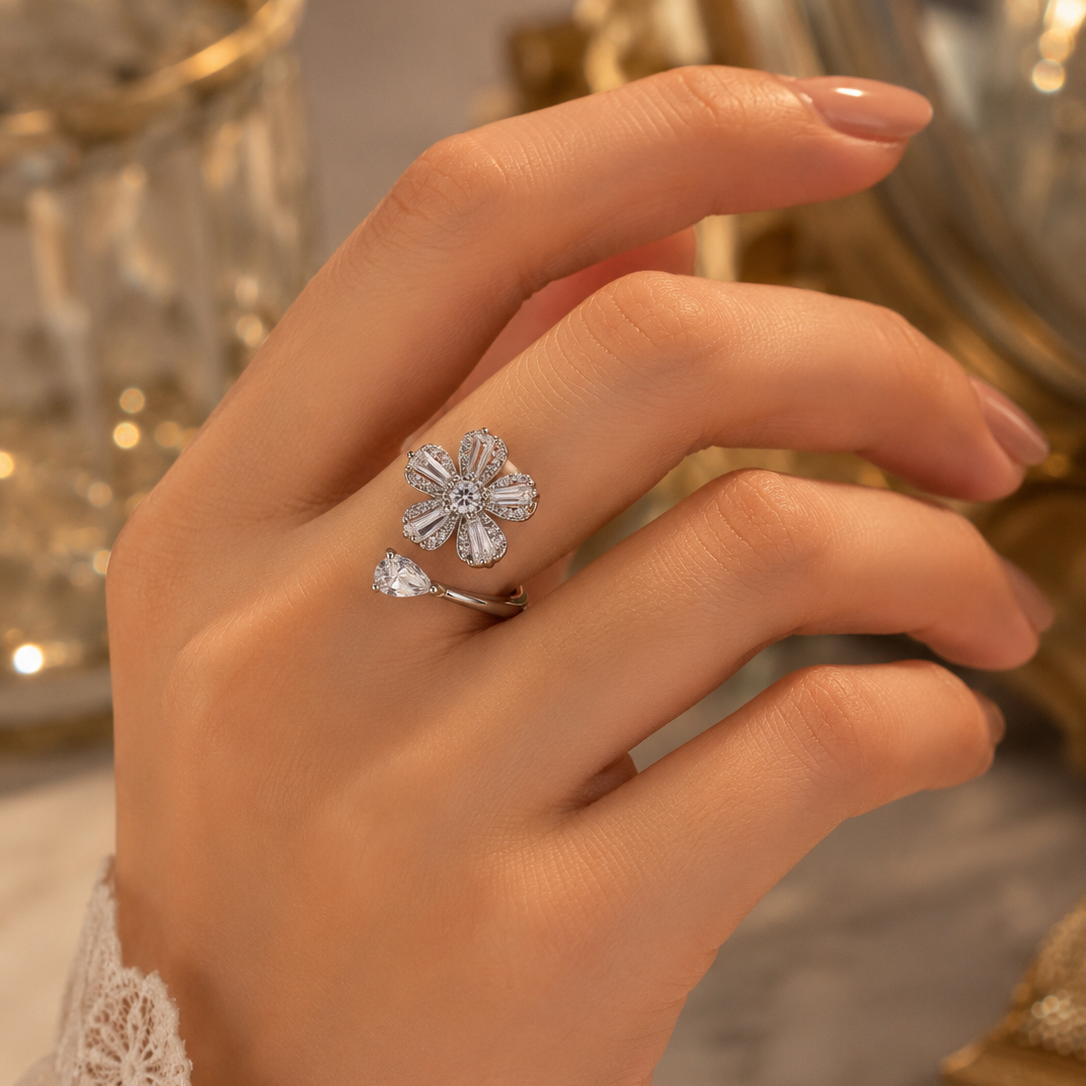 A silver flower-shaped ring with a dangling teardrop charm, worn on a woman's hand against a warm, softly blurred background.