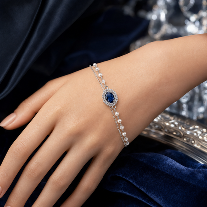 A close-up of a woman's hand wearing a delicate silver-toned bracelet with a central oval blue gemstone surrounded by small clear stones, accented by rows of white pearls on the chain.