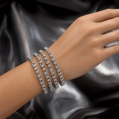 Four slim silver tennis bracelets, each set with clear round-cut stones, stacked on a woman's wrist against a dark satin background.