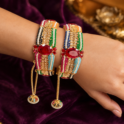 A close-up of a person's wrist and hand wearing two ornate gold-plated bangles decorated with colorful beaded stripes and large red oval stones, with dangling chain tassels resting against a deep purple velvet fabric.
