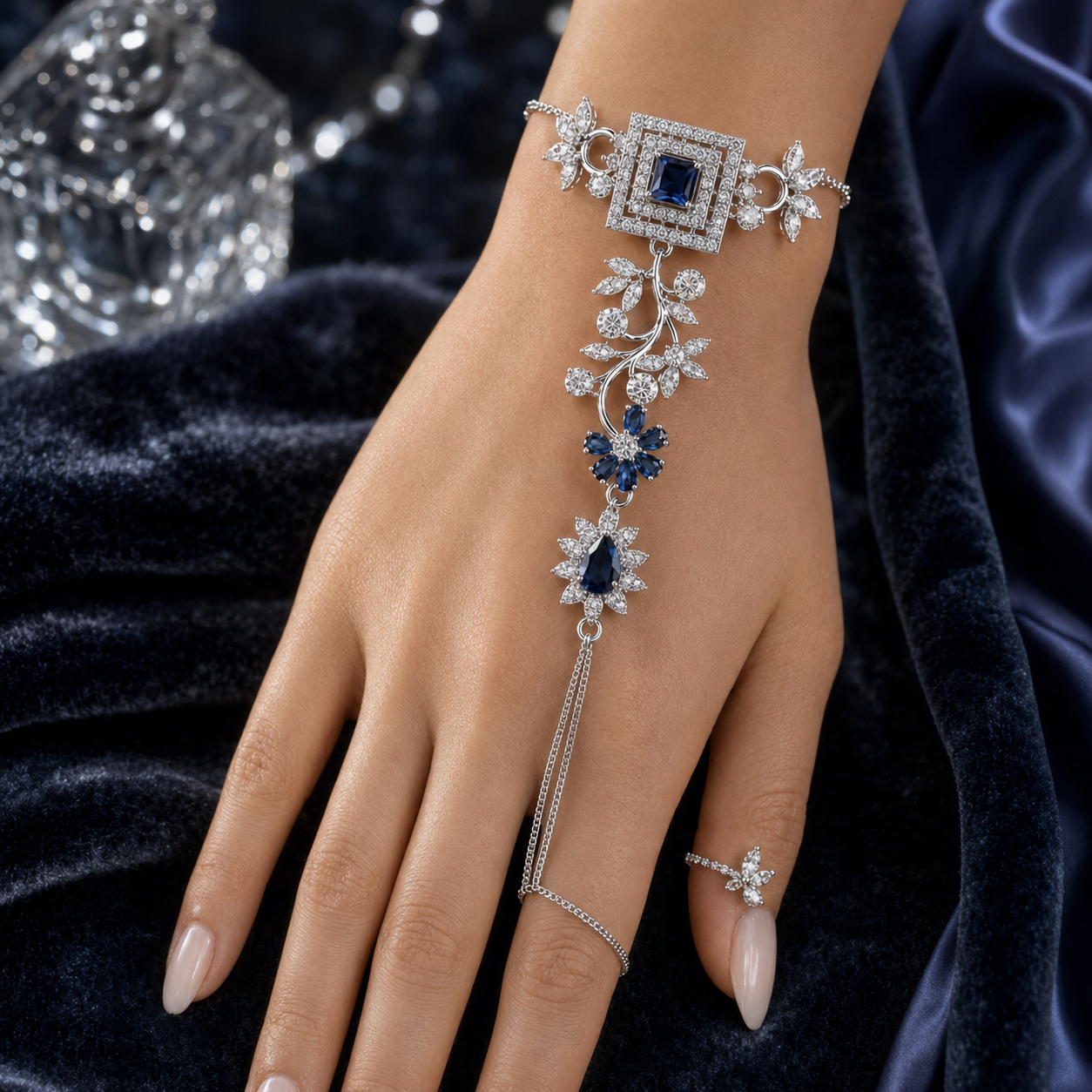 A close-up of a hand wearing an ornate silver-toned hand chain bracelet with deep blue gemstones and clear crystal-like stones, styled against dark velvet fabric.