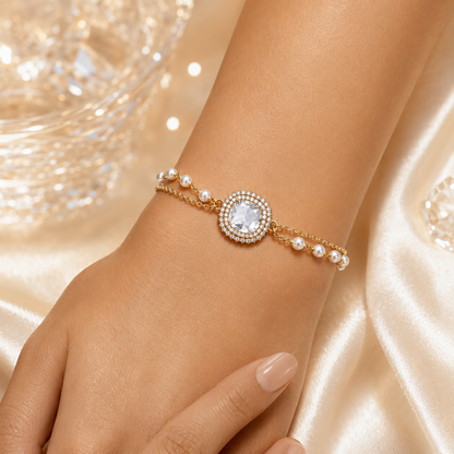 A gold-toned double-strand bracelet with a central round clear gemstone surrounded by small pearls, shown worn on a person's wrist against a soft champagne satin background.