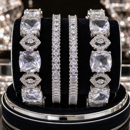 Four silver-toned diamond-like jewelry pieces displayed on a black velvet ring stand: two wide bands with large cushion-cut clear stones and two slim bands with pavé-set clear stones, photographed against a sparkling reflective background.