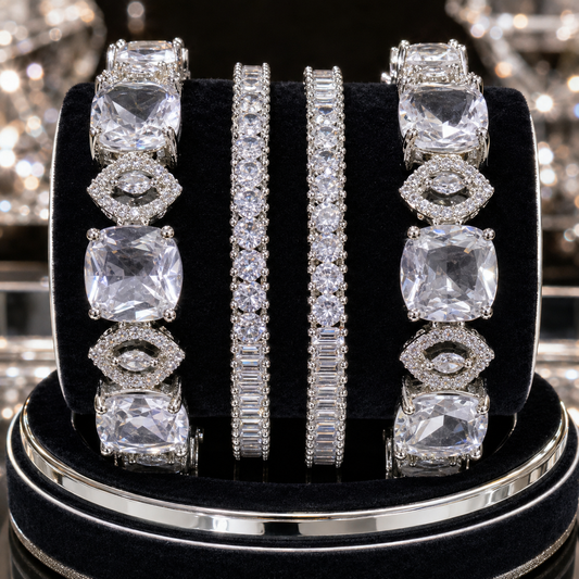 Four silver-toned diamond-like jewelry pieces displayed on a black velvet ring stand: two wide bands with large cushion-cut clear stones and two slim bands with pavé-set clear stones, photographed against a sparkling reflective background.