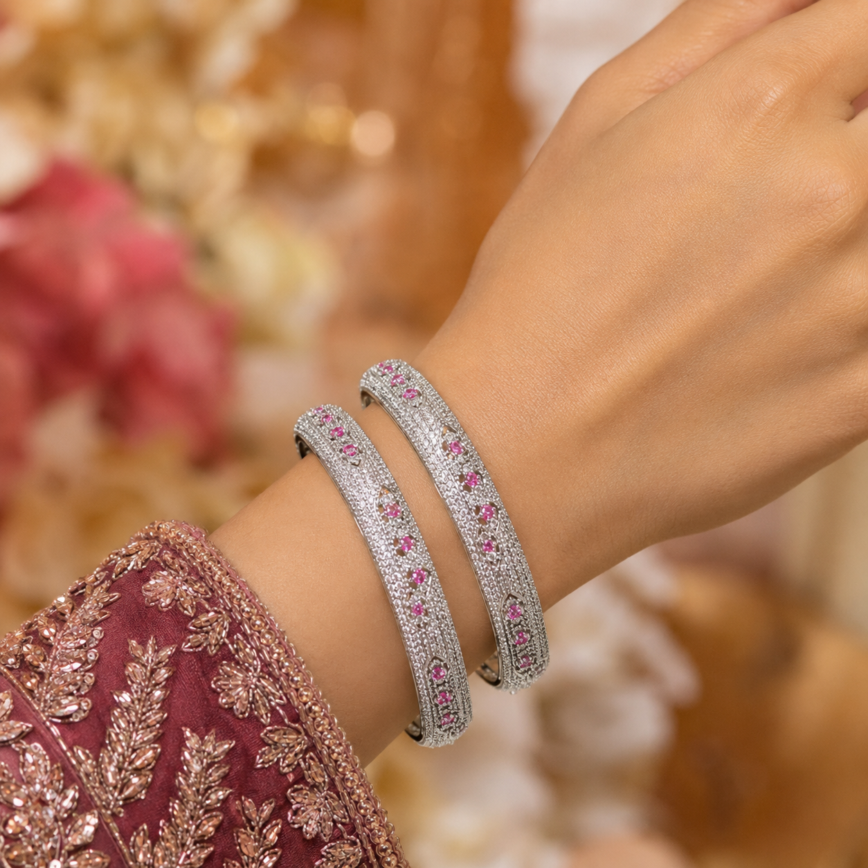 A close-up of a person's wrist wearing two silver-toned bangles adorned with small pink stones, set against a softly blurred warm-toned background.
