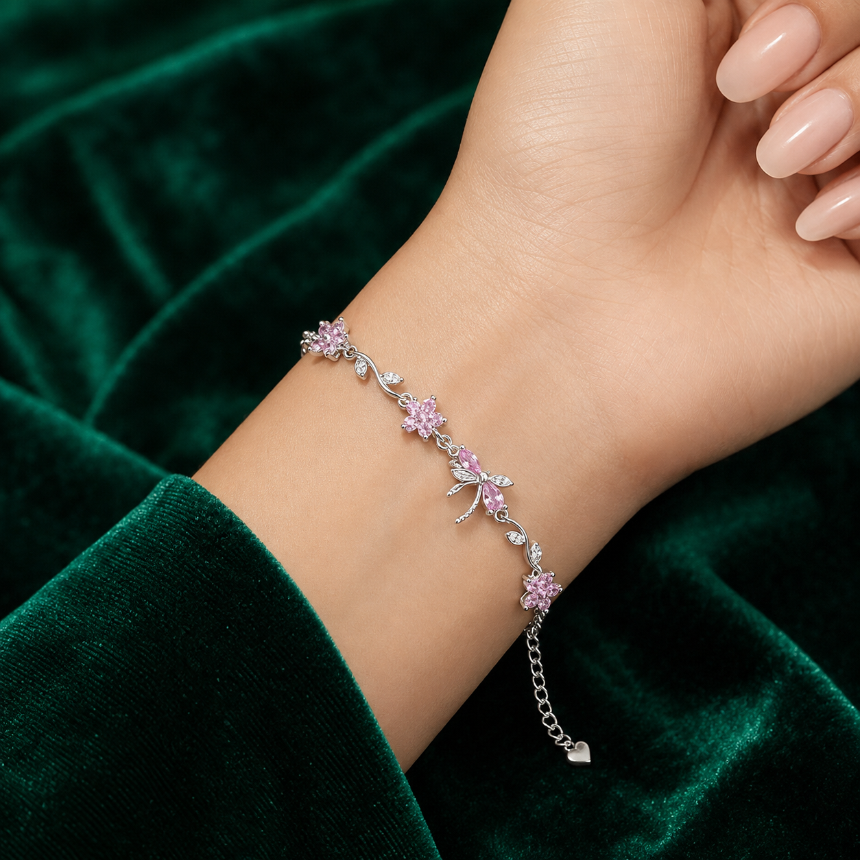 A delicate silver chain bracelet with pink flower and butterfly motifs, worn on a woman's wrist against a dark green velvet background.