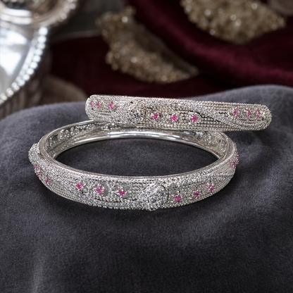 A silver-toned bangle bracelet with intricate pavé detailing and small pink gemstone accents, displayed on a dark gray velvet surface.