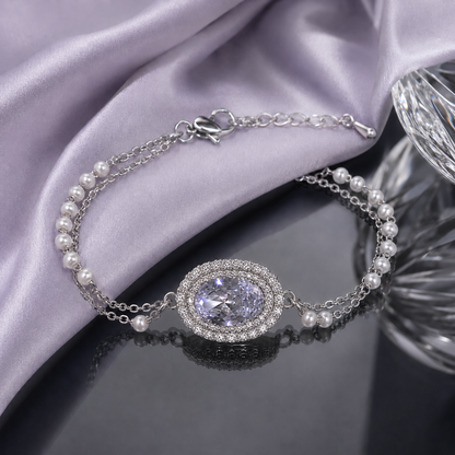 A silver chain bracelet with white pearls and a central oval crystal pendant surrounded by a halo of small clear stones, displayed on draped lavender satin fabric over a dark reflective surface.