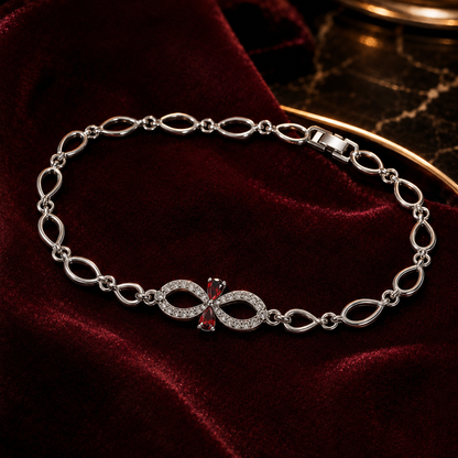 A silver-toned link bracelet with an infinity symbol centerpiece, accented by a small red gemstone and clear stones, displayed on a dark red velvet surface.